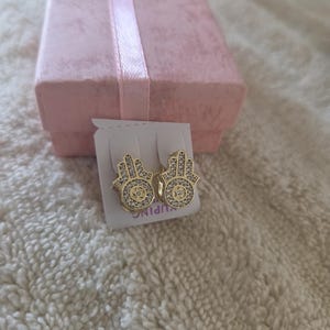 Puede incluir: Un par de pendientes dorados con forma de mano de Hamsa, con piedras claras y brillantes. Los pendientes se muestran en una tarjeta blanca, con una caja de regalo rosa en el fondo. Los pendientes son un símbolo de protección y buena suerte.