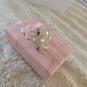 Puede incluir: Un anillo plateado con un grupo de piedras ovaladas, cada una rodeada de piedras más pequeñas y transparentes. El anillo está sobre una caja de regalo rosa atada con una cinta blanca, sobre una superficie texturizada blanquecina.