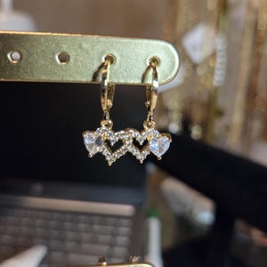 Puede incluir: Pendientes dorados con tres diseños en forma de corazón, cada uno adornado con piedras preciosas claras y brillantes. Los pendientes tienen un cierre de palanca y se exhiben en un soporte dorado. El diseño es delicado y elegante.