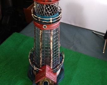 May include: A red, white, and blue metal lighthouse figurine with a glass window and a wire mesh top. The lighthouse is sitting on a green surface.