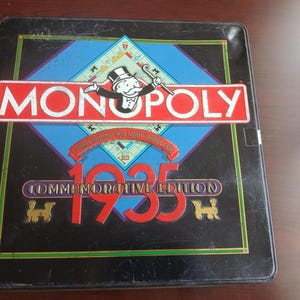 May include: A vintage Monopoly Commemorative Edition board game from 1935. The game box is black with a red and white Monopoly logo, a blue diamond, and the number 1935 in red. The box has a gold border.