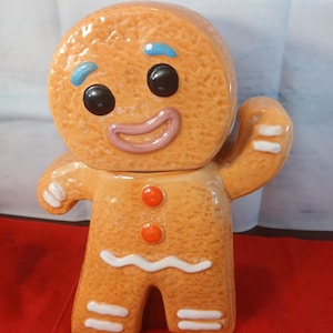 Puede incluir: Un tarro de galletas de cerámica con forma de hombre de jengibre, con superficie texturizada de color tostado, ojos negros, cejas azules y boca rosa. Presenta dos botones naranjas en el pecho y detalles de glaseado blanco en brazos y piernas.