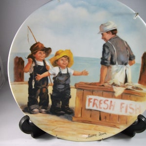 Puede incluir: Un plato de cerámica con una pintura de dos niños pescando y un hombre vendiendo pescado fresco. Los niños llevan monos y sombreros. El hombre lleva un delantal blanco y un sombrero blanco. El plato tiene un fondo azul y blanco con un muelle de madera y un cartel que dice "Pescado fresco".