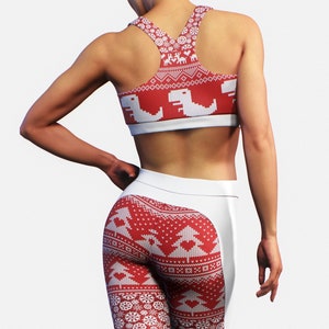 Op de afbeelding: Rode en witte sportset met kerstthema. De sport-bh en legging hebben een feestelijk patroon met dinosaurussen, bomen, sneeuwvlokken en rendieren. De legging heeft een wit paneel aan de zijkanten.
