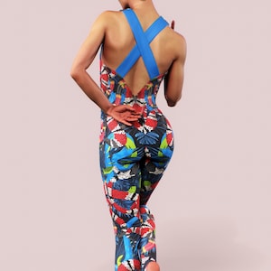 Könnte beinhalten: Ein einteiliger Jumpsuit mit einem lebendigen tropischen Druck, der rote, blaue und grüne Elemente auf dunklem Hintergrund aufweist. Der Jumpsuit hat ein gekreuztes Rückendesign mit blauen Trägern.