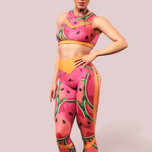 Puede incluir: Conjunto deportivo rosa con estampado de sandía. El conjunto incluye un sujetador deportivo y leggings. Los leggings y el sujetador tienen una base rosa con rodajas de sandía en rojo, verde y negro. También hay detalles en naranja.