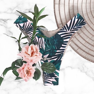 Op de afbeelding: Hoog getailleerde bikinibroek met een tropische bladerprint in groen en wit. De broek is gecombineerd met roze rozen en bamboestengels. Op de achtergrond is een traditionele conische hoed te zien.
