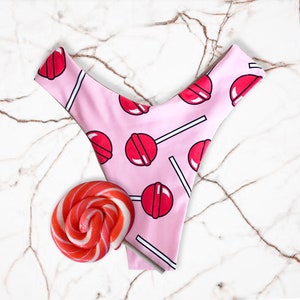 May include: Pink underwear with a lollipop pattern, featuring red lollipops on white sticks. A red and white swirled lollipop is next to the underwear on a marble surface. The underwear has a V-shaped front.