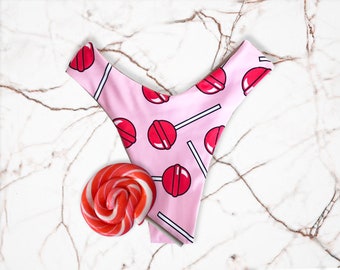 May include: Pink underwear with a lollipop pattern, featuring red lollipops on white sticks. A red and white swirled lollipop is next to the underwear on a marble surface. The underwear has a V-shaped front.