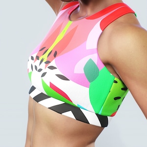 Puede incluir: Un sujetador deportivo colorido con un vibrante estampado floral. El sujetador presenta una mezcla de rosa, verde, rojo y blanco, con acentos geométricos en blanco y negro en la parte inferior. El diseño es moderno y llamativo.