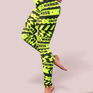 May include: Neon yellow leggings with black warning tape design. The leggings feature the words "WARNING", "CAUTION", and "DO NOT CROSS" in black. The leggings have a high waist and a form-fitting design.