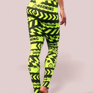 May include: Neon green leggings with a black hazard stripe pattern and the words "WARNING" and "CAUTION" printed throughout. The leggings have a high waist and a matching neon green waistband.