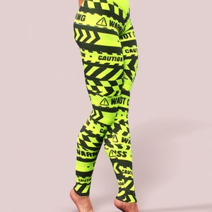 May include: Neon yellow leggings with a black warning tape design. The leggings feature the words "WARNING" and "CAUTION" in black, along with hazard symbols. The high-waisted leggings are made of a stretchy material.