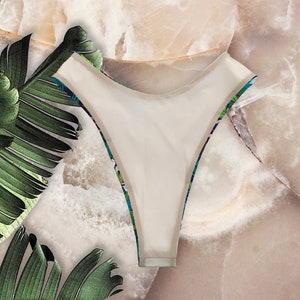 May include: White swimsuit with a tropical print. The swimsuit has a high-cut leg and a low back.