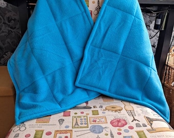 May include: A vibrant turquoise quilted fleece wrap, approximately 150cm long, draped over a chair. The wrap features a diamond pattern and is made of soft fleece. The chair has a patterned fabric with sewing-related images. Designed to be worn around the neck and shoulders.