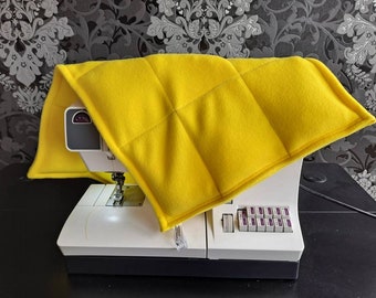 May include: A vibrant yellow, quilted sewing machine cover resting on a white and grey sewing machine. The cover is rectangular with a diamond pattern and is made of a soft material. The sewing machine has a needle, a light, and several buttons.