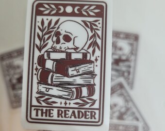 May include: A white sticker with a brown border and a skull sitting on top of a stack of books. The sticker has the text "THE READER" at the bottom.
