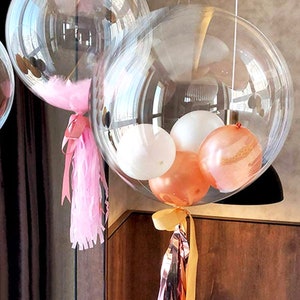 May include: A large clear balloon filled with smaller balloons in white, peach, and rose gold. The balloon is tied with a pink tassel and a gold ribbon.