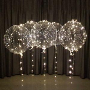 May include: A set of four clear plastic balloons with white fairy lights inside. The balloons are hanging from the ceiling and the lights are cascading down.