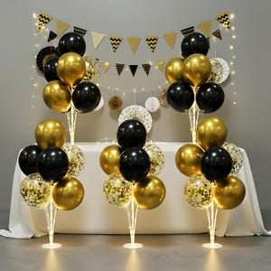 May include: A festive party decoration set featuring black, gold, and confetti balloons arranged on illuminated stands. The backdrop includes a banner with gold and black chevron and triangle flags, along with string lights and paper fans.