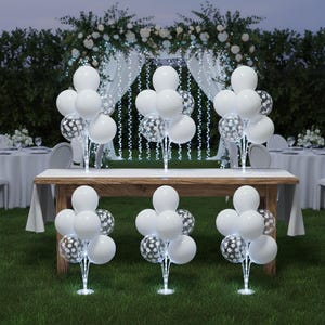 May include: White and confetti-filled balloons arranged in clusters on clear stands, illuminated with small lights. The balloons are set up on a wooden table and on the grass, with a wedding arch and tables in the background.