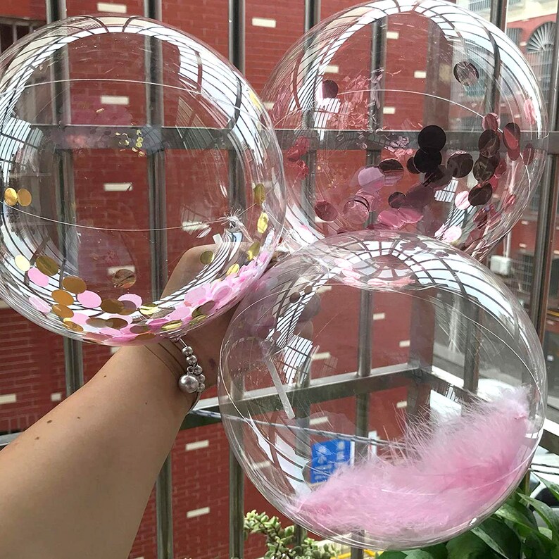 May include: Three clear plastic balloons filled with confetti and feathers. The balloons are tied together and held by a hand. The confetti is pink and gold, and the feathers are pink.
