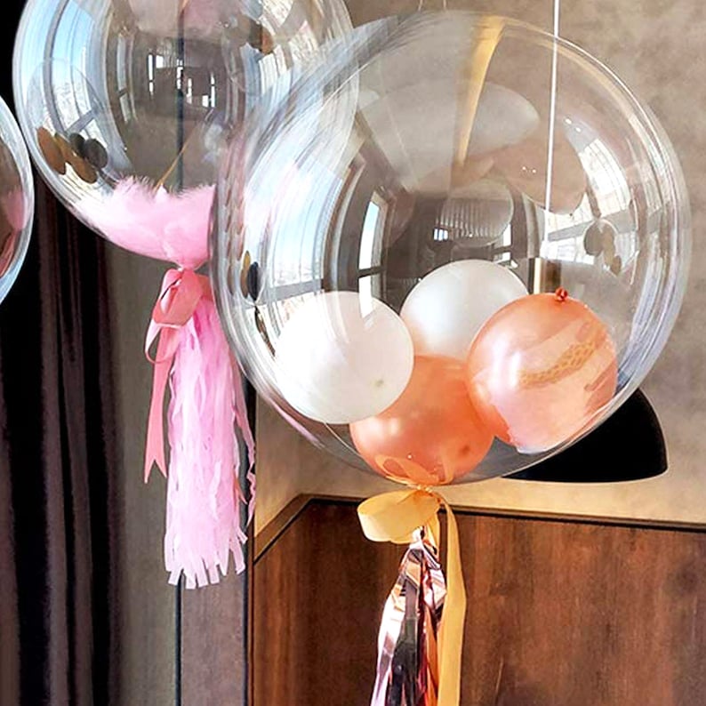 Pu&ograve; includere: Un grande palloncino trasparente riempito con palloncini pi&ugrave; piccoli in bianco, pesca e oro rosa. Il palloncino &egrave; legato con un fiocco rosa e un nastro dorato.