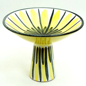 May include: A yellow and black ceramic pedestal bowl with a geometric design. The bowl is shaped like a flower with a wide, shallow base and a tall, narrow stem.