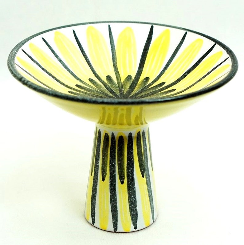 May include: A yellow and black ceramic pedestal bowl with a geometric design. The bowl is shaped like a flower with a wide, shallow base and a tall, narrow stem.
