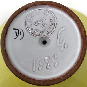 May include: Close-up of the bottom of a white ceramic vase with a brown rim. The vase has a brown stamp that reads "Laholm Made in Sweden" and the number "1395" written in white.