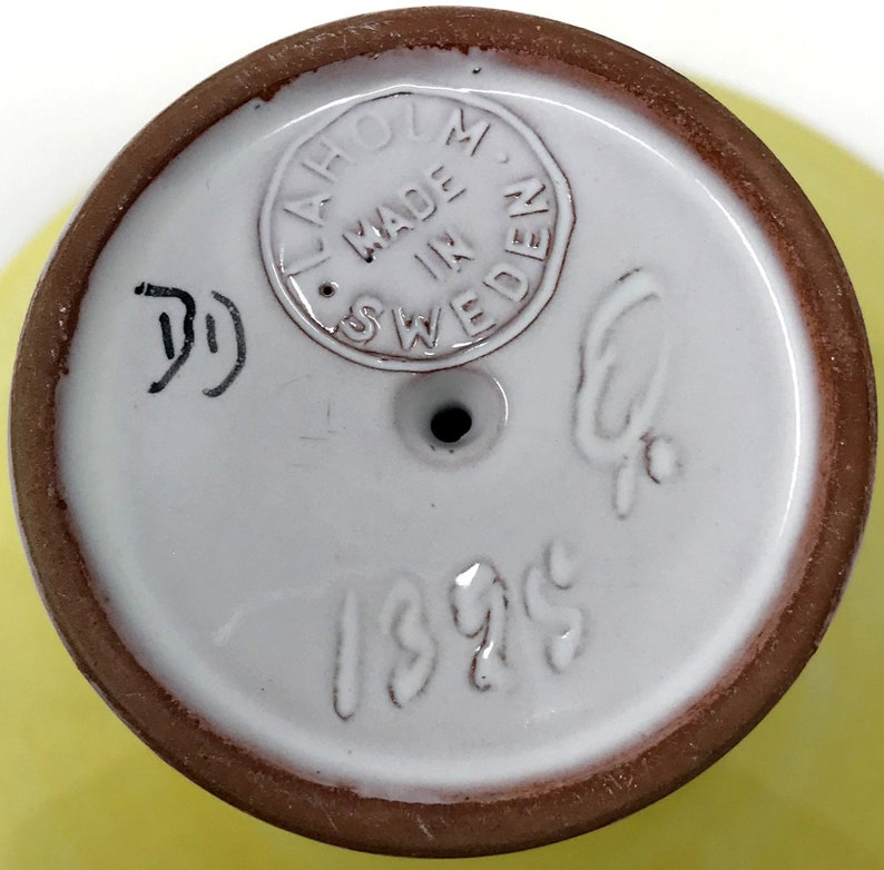 May include: Close-up of the bottom of a white ceramic vase with a brown rim. The vase has a brown stamp that reads "Laholm Made in Sweden" and the number "1395" written in white.