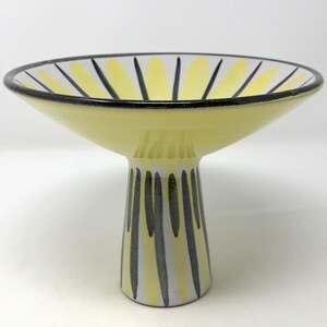 May include: A yellow ceramic pedestal bowl with black and white stripes. The bowl is wide and shallow with a tall, narrow base.