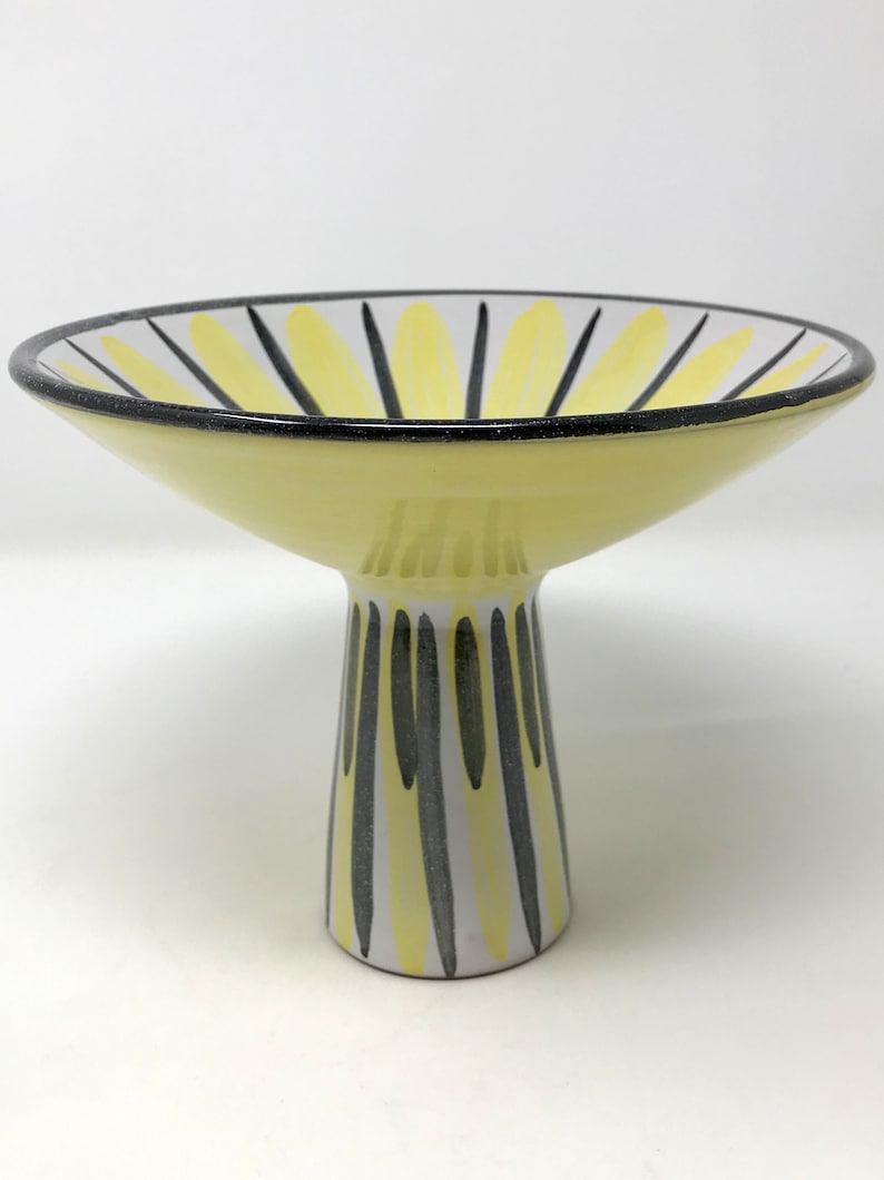 May include: A yellow ceramic pedestal bowl with black and white stripes. The bowl is wide and shallow with a tall, narrow base.