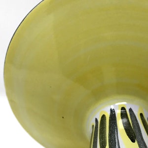 May include: A yellow ceramic cake stand with a white base and black stripes. The stand has a wide, flat top and a tall, narrow base.
