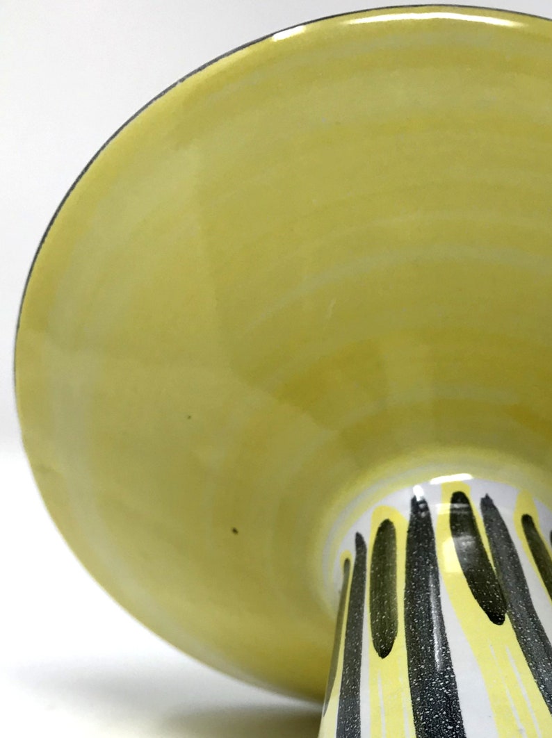 May include: A yellow ceramic cake stand with a white base and black stripes. The stand has a wide, flat top and a tall, narrow base.