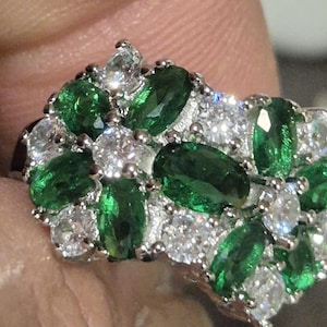May include: A cluster ring featuring emerald-green and clear gemstones set in a silver-toned band. The ring's design showcases a mix of oval and round-cut stones, creating a sparkling, eye-catching piece of jewelry.