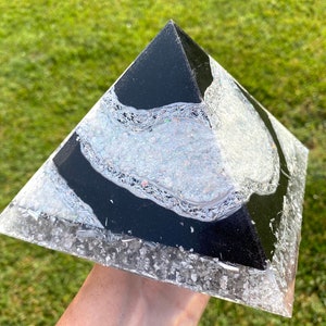 May include: A handcrafted pyramid with a black top and a clear base. The pyramid features a design with silver glitter and a black stripe. The pyramid is held in a hand against a green grassy background.