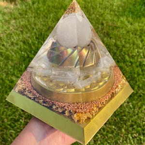 May include: A handcrafted pyramid-shaped art piece with a gold-colored base and accents. The pyramid features a clear resin exterior, with a white sphere at the top and gold and iridescent elements inside. The base is square, with a gold border.