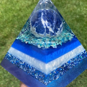 May include: A layered pyramid sculpture with a blue and white color scheme. A sphere of blue and white stone sits inside the pyramid. The pyramid is made of resin and has layers of blue, white, and glittery accents. The sculpture is held in a hand.