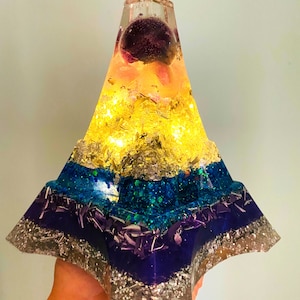 May include: A multi-layered resin pyramid with a clear top and a purple crystal sphere inside. The pyramid is decorated with gold flakes, blue glitter, and silver glitter.