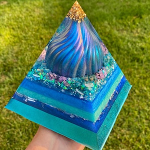May include: A handcrafted pyramid sculpture with layers of teal, turquoise, and blue resin. The top features a swirling blue design and is topped with gold leaf. Decorative crystals and glitter are embedded in the resin.