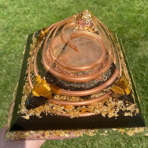 May include: A handcrafted pyramid-shaped object with a clear resin exterior. The pyramid features copper coils, gold flakes, and crystals. The base is black with gold leaf accents, and the top is adorned with crystals.