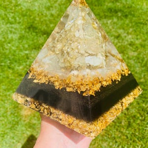 May include: A pyramid-shaped decorative object with a clear resin exterior. The pyramid is filled with gold flakes and a black band near the base. The pyramid is held in a hand against a green background.