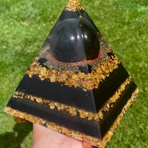 May include: A black pyramid-shaped resin art piece with gold leaf accents and a black sphere on top. The pyramid is held in a hand against a backdrop of green grass.