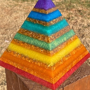 May include: A rainbow-colored pyramid made of resin with gold glitter embedded in each layer. The pyramid is sitting on a wooden surface.