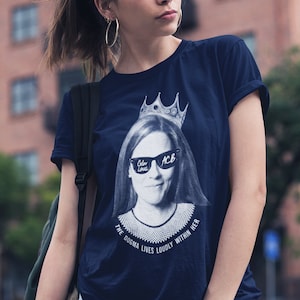 May include: A navy blue t-shirt with a white graphic of Ruth Bader Ginsburg wearing sunglasses and a crown. The text "The dogma lives loudly within her" is printed below the image.