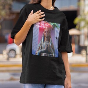 May include: Black t-shirt featuring a graphic print of a woman in a blue robe with the text "I'm not like other girls" in pink and white.