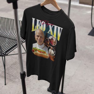 K&ouml;nnte beinhalten: Schwarzes T-Shirt mit einer Grafik eines Mannes in religi&ouml;ser Kleidung, der einen Hotdog h&auml;lt. Der Text "LEO XIV" ist in Gelb und Wei&szlig; dargestellt, mit dem Wort "Pope" in Rot dar&uuml;ber. Zwei kleinere Bilder sind ebenfalls enthalten.