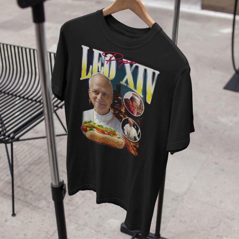 K&ouml;nnte beinhalten: Schwarzes T-Shirt mit einer Grafik eines Mannes in religi&ouml;ser Kleidung, der einen Hotdog h&auml;lt. Der Text "LEO XIV" ist in Gelb und Wei&szlig; dargestellt, mit dem Wort "Pope" in Rot dar&uuml;ber. Zwei kleinere Bilder sind ebenfalls enthalten.