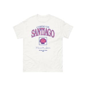 May include: White t-shirt with a blue and pink graphic design that says "Camino de Santiago" with a scallop shell and the text "France to Spain, The Way of St. James 1982".